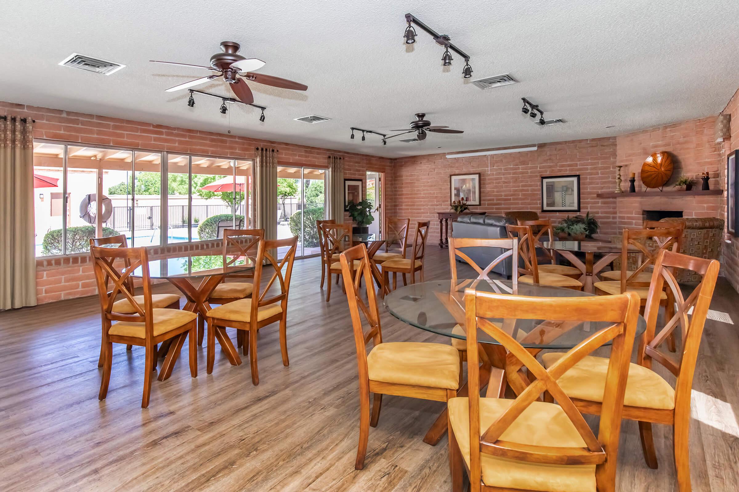 A spacious indoor gathering area featuring several wooden tables with chairs, large windows allowing natural light, and a warm ambiance created by brick walls. Ceiling fans and track lighting enhance the comfort of the space, which is suitable for events or social gatherings.
