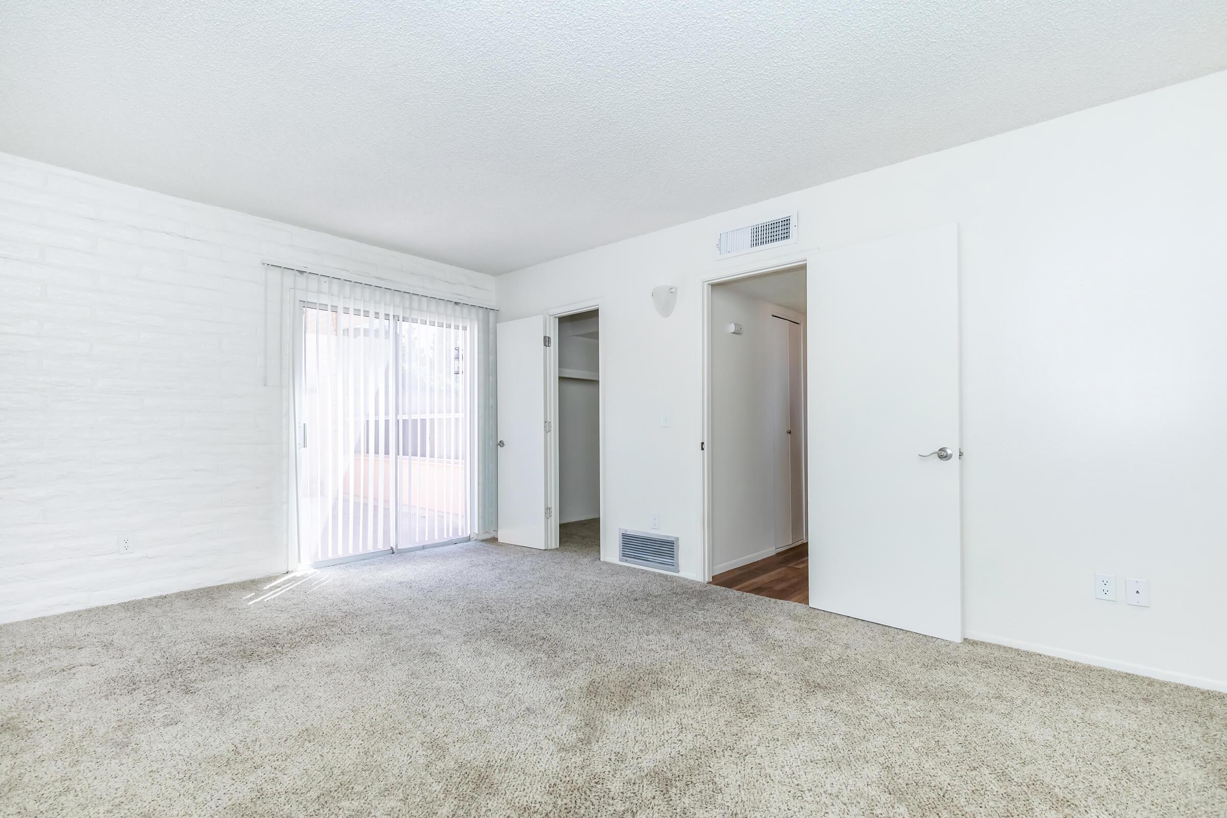 A spacious, empty room featuring light-colored walls, a textured white accent wall, and plush beige carpeting. There are sliding glass doors leading to an outdoor area, as well as two doorways leading to other rooms. Natural light brightens the space, creating an inviting atmosphere.